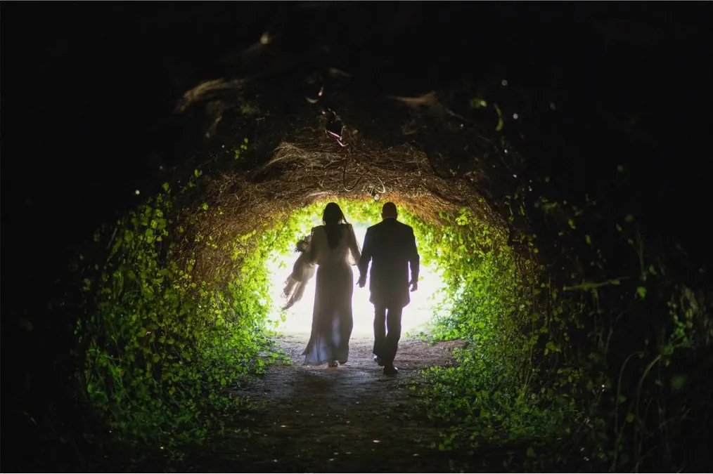 Novios caminando bajo un túnel de vegetación hacia la luz